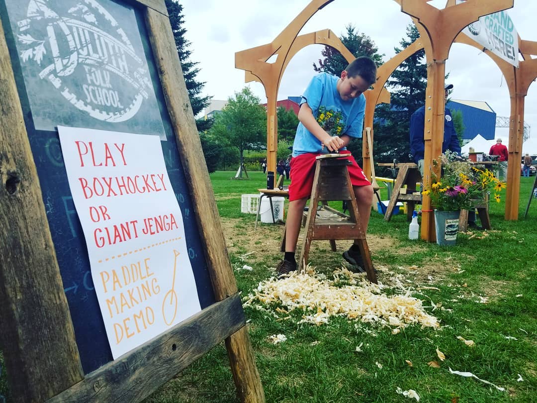 Lake Superior Harvest Festival at Bayfront in Duluth, with a young kid carving a paddle with shavings on the ground.