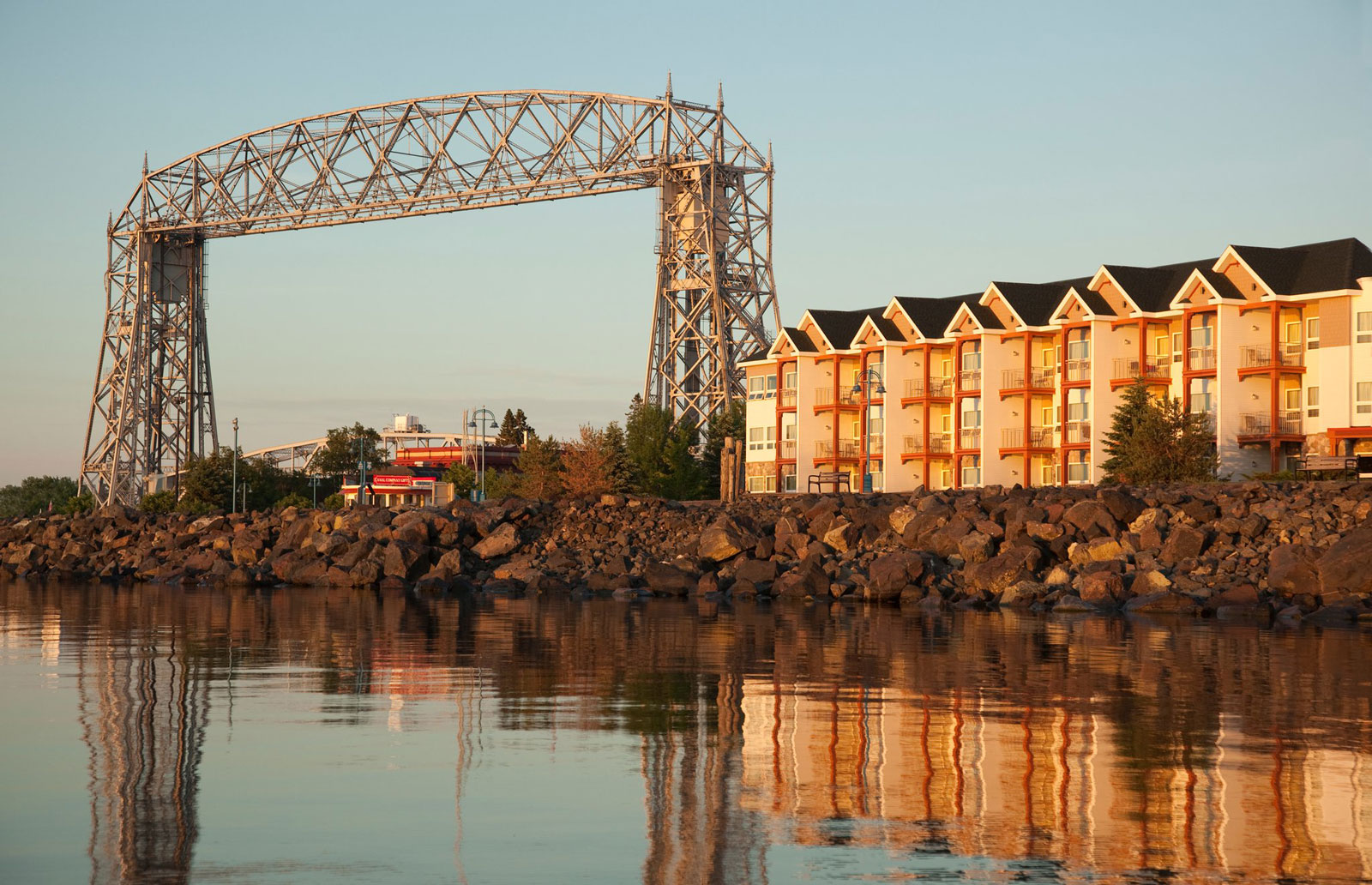 A view of the Aerial Lift Bridge in Duluth, MN with the sunrise hitting the glass-like water and the rocky shoreline.