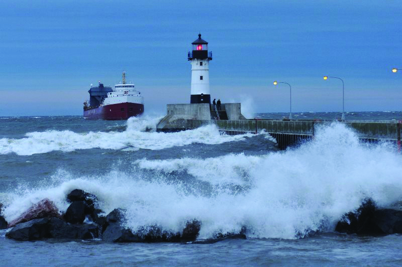 The waves of Lake Superior crashing into the shoreline and the Lighthouse with a ship entering the canal.