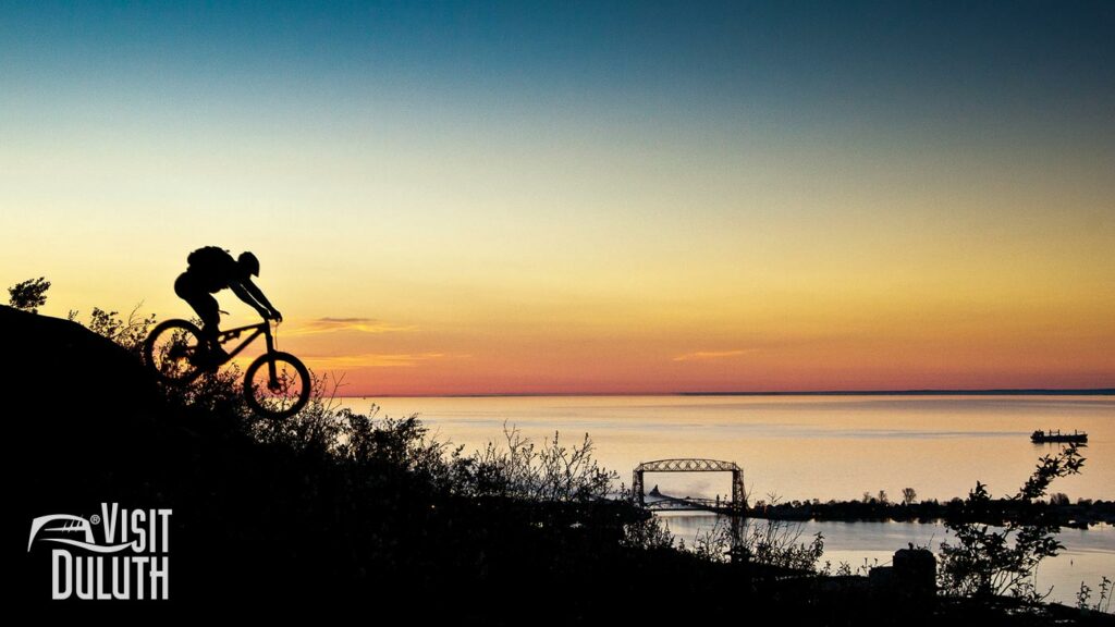 A silhouetted mountain biker going down a hill during sunset. The Aerial Lift Bridge and a ship are in the distance.