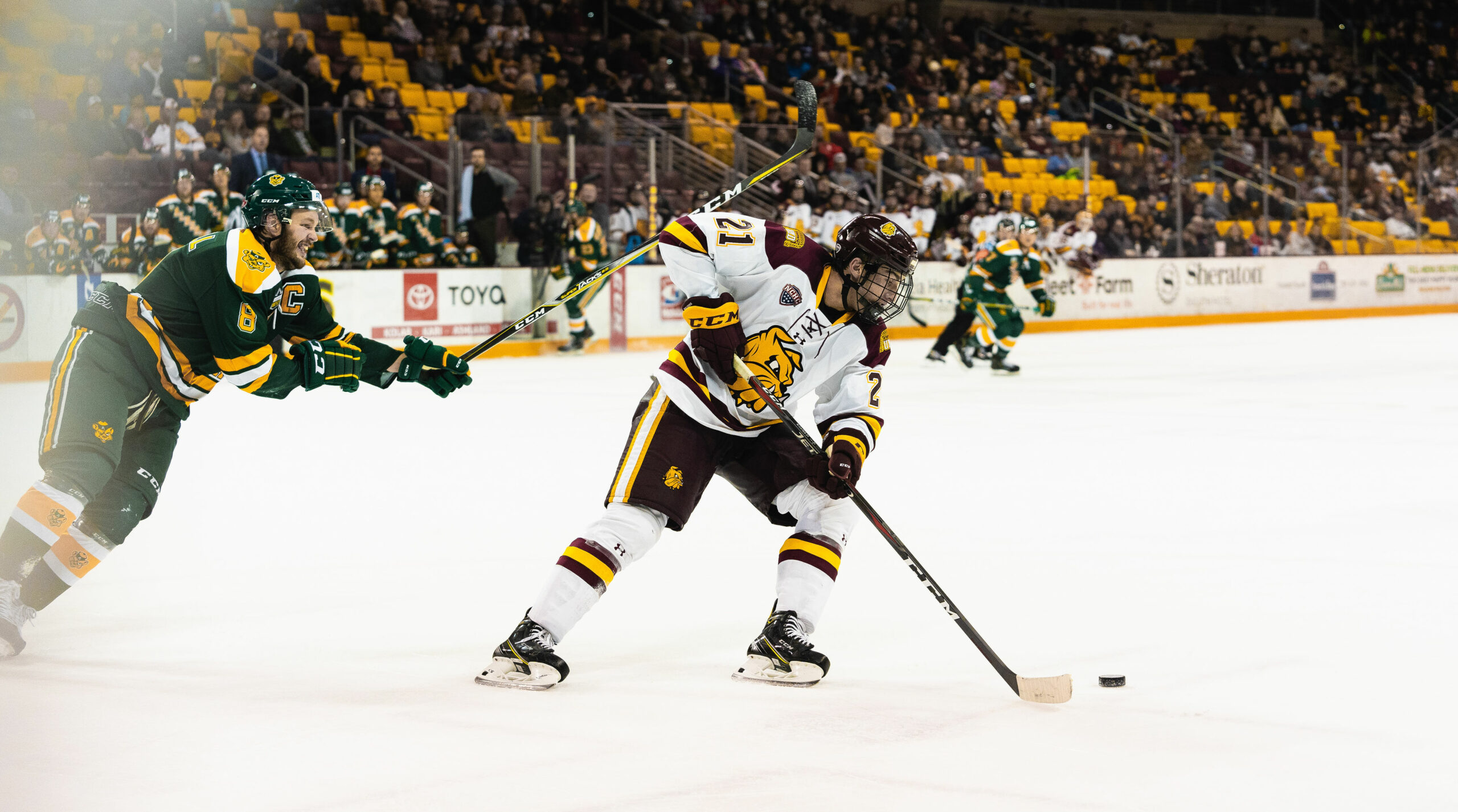 UMD Mens hockey game. A player is chasing the puck and another player is hitting him in the shoulder with his stick.