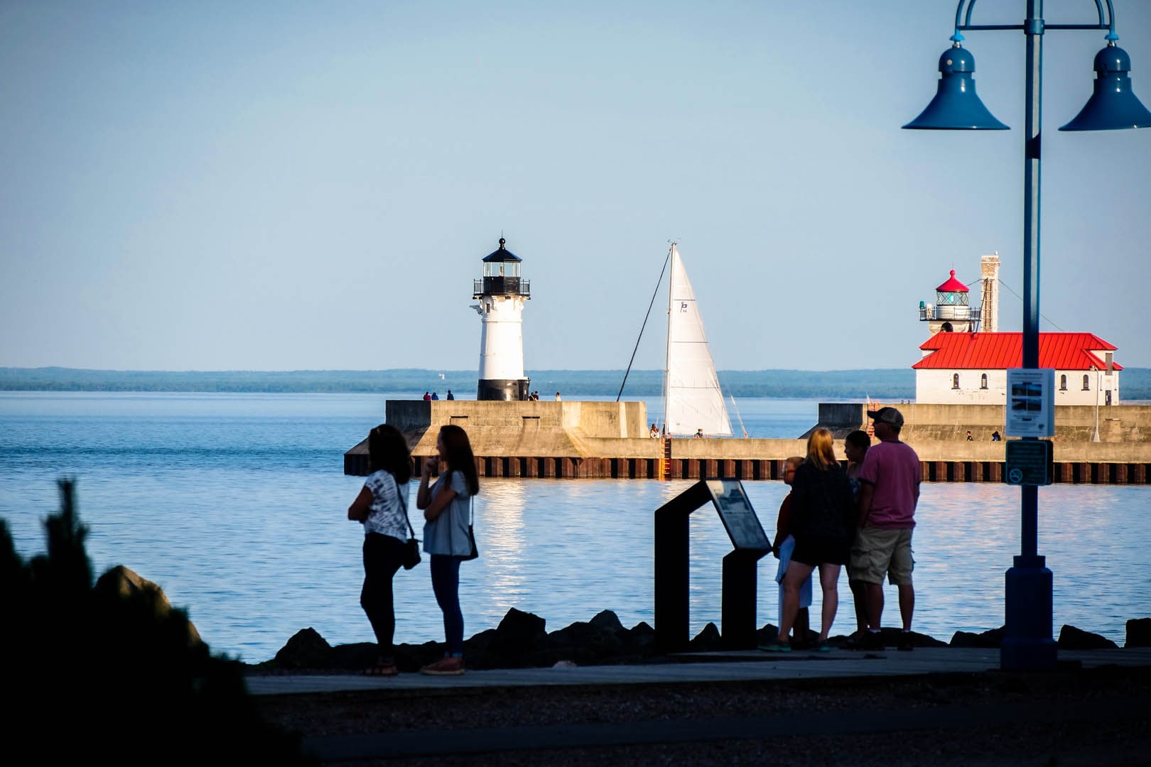 Two groups of people in silhouette looking out at very blue water with the piers and lighthouses behind in full sun.