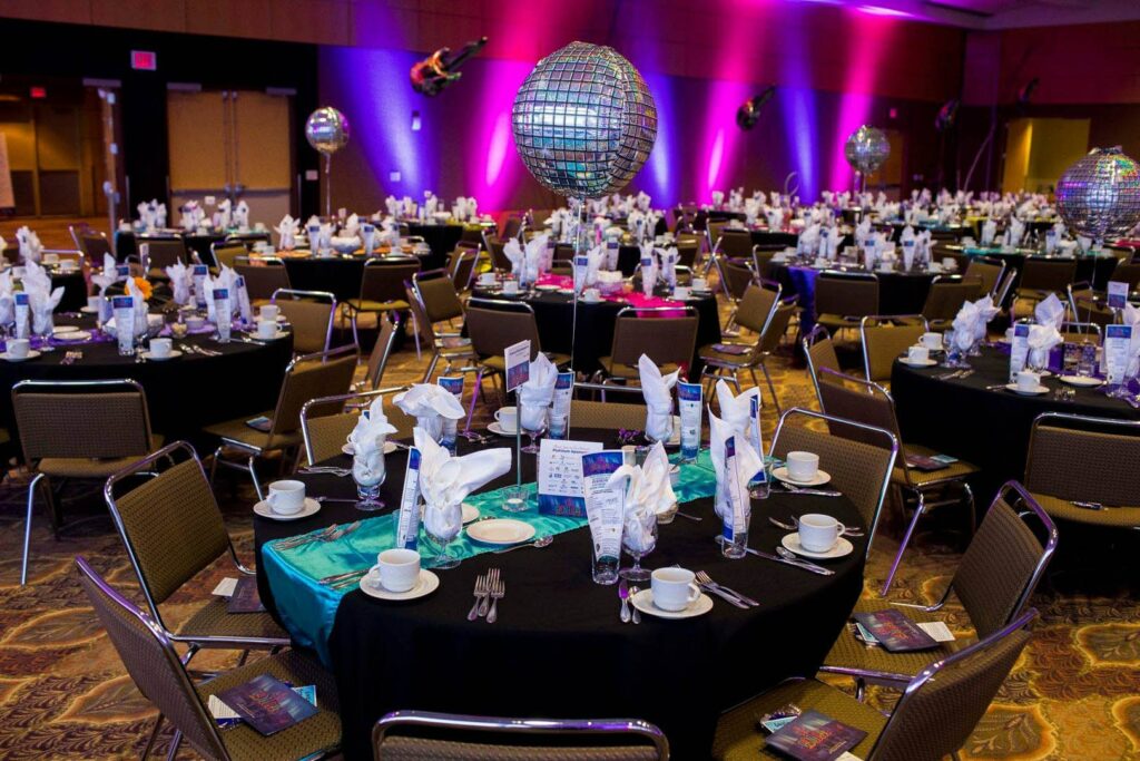 Event space with twenty tables set, bright party lights flank the wall and silver helium balloons are at every table.