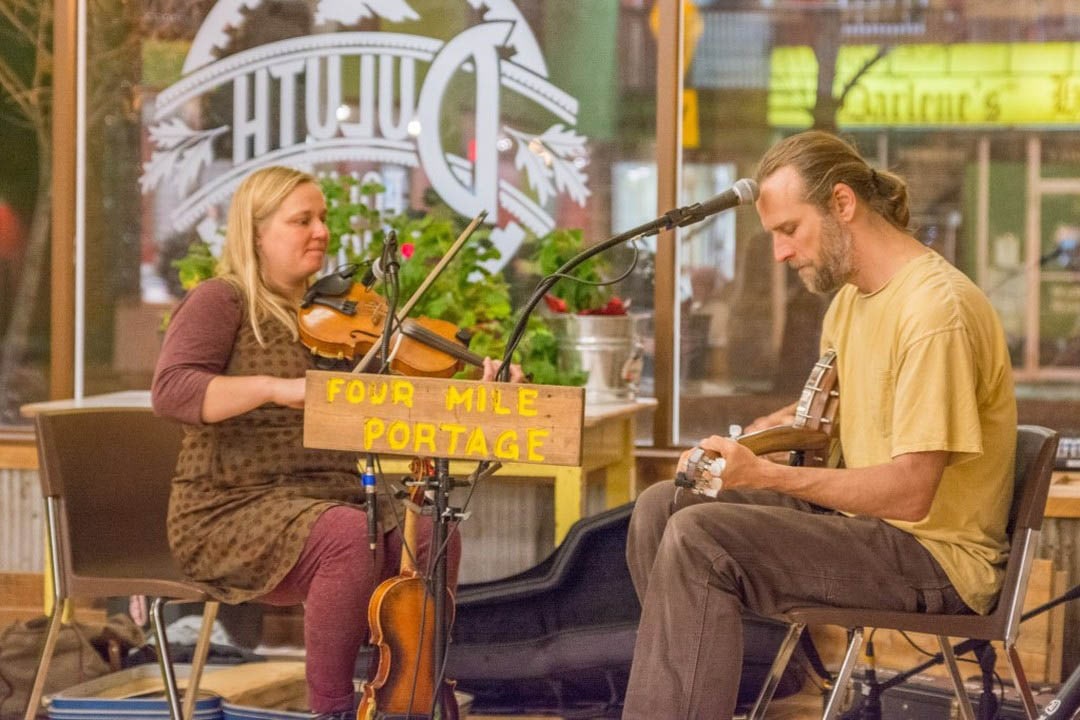 Man and woman sitting in chairs, she is playing violin and he is playing a banjo.