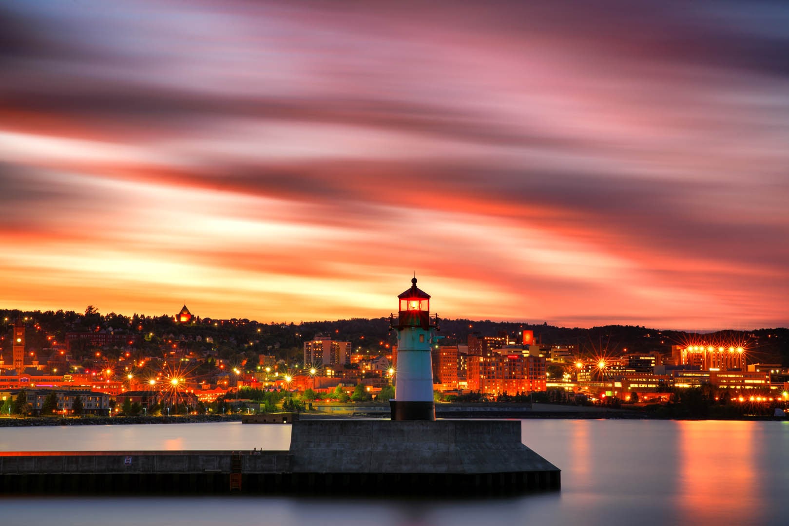 Orange sunset with lighthouse in the foreground and city lights in the distance.
