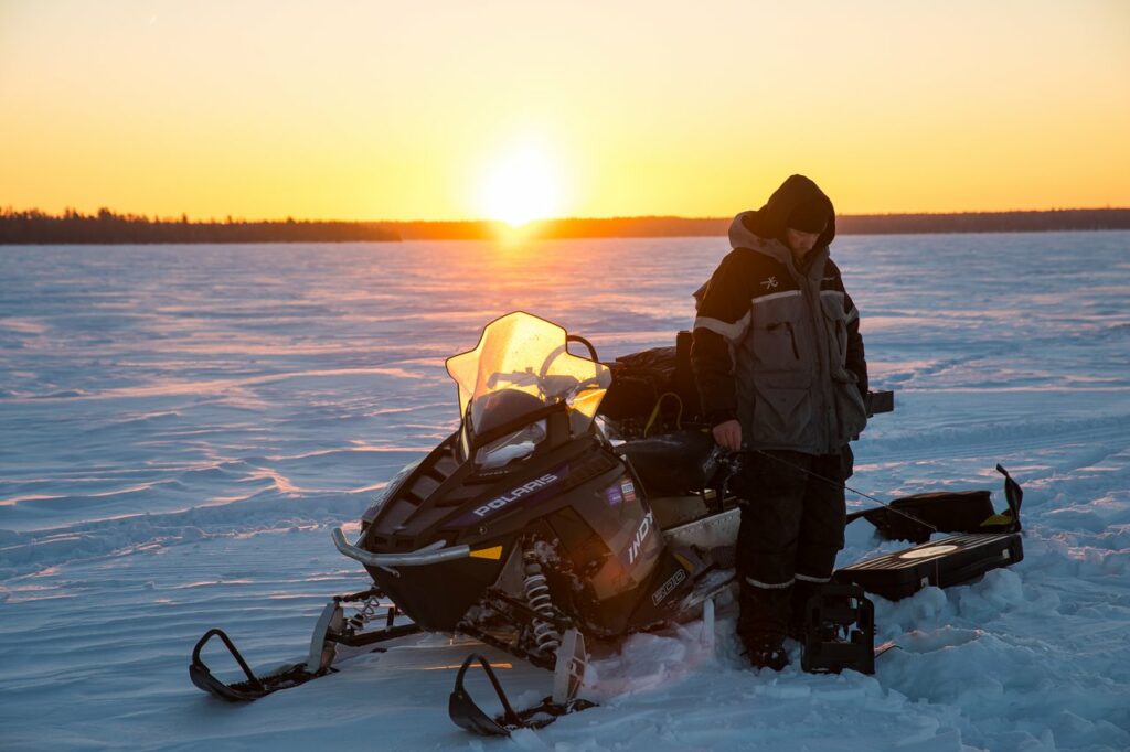 A man in winter gear standing next to his snowmobile on a frozen lake. He is ice fishing.