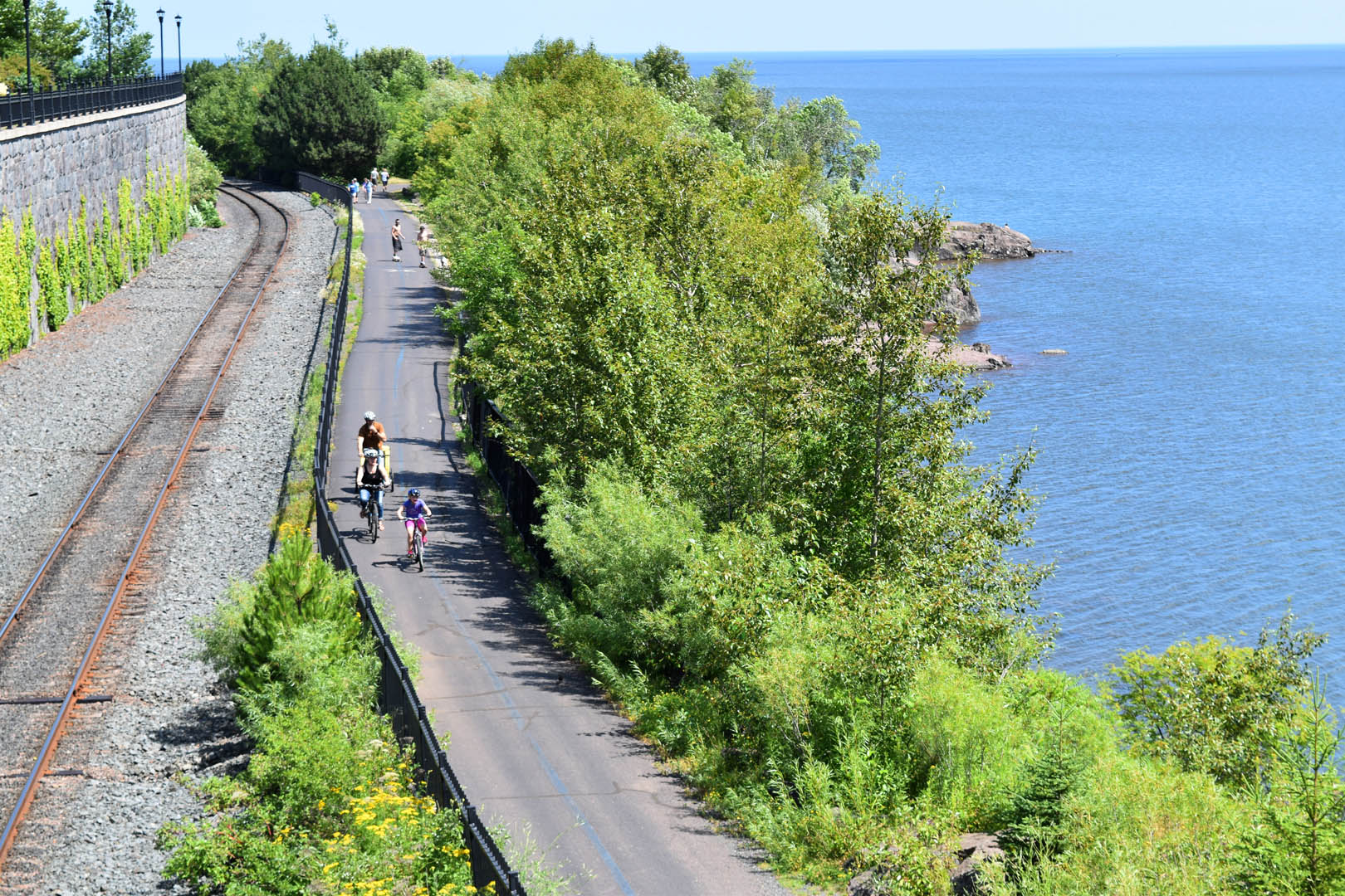 Looking down from above at bikers and walkers on the Duluth Lakewalk with green trees and a very blue lake superior