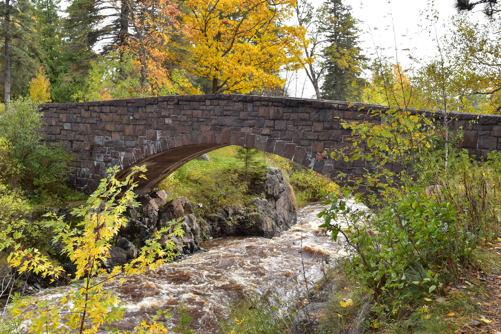Looking up at an arched bridge along a rapidly flowing Lester River in autumn.