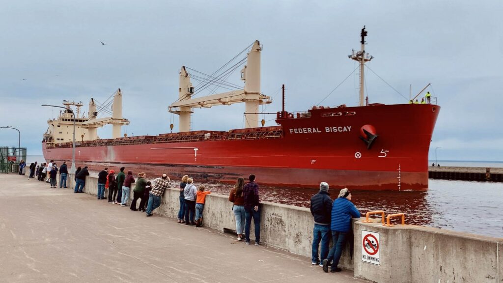 People watch a huge red ship go through the canal. The ship's name is Federal Biscay.