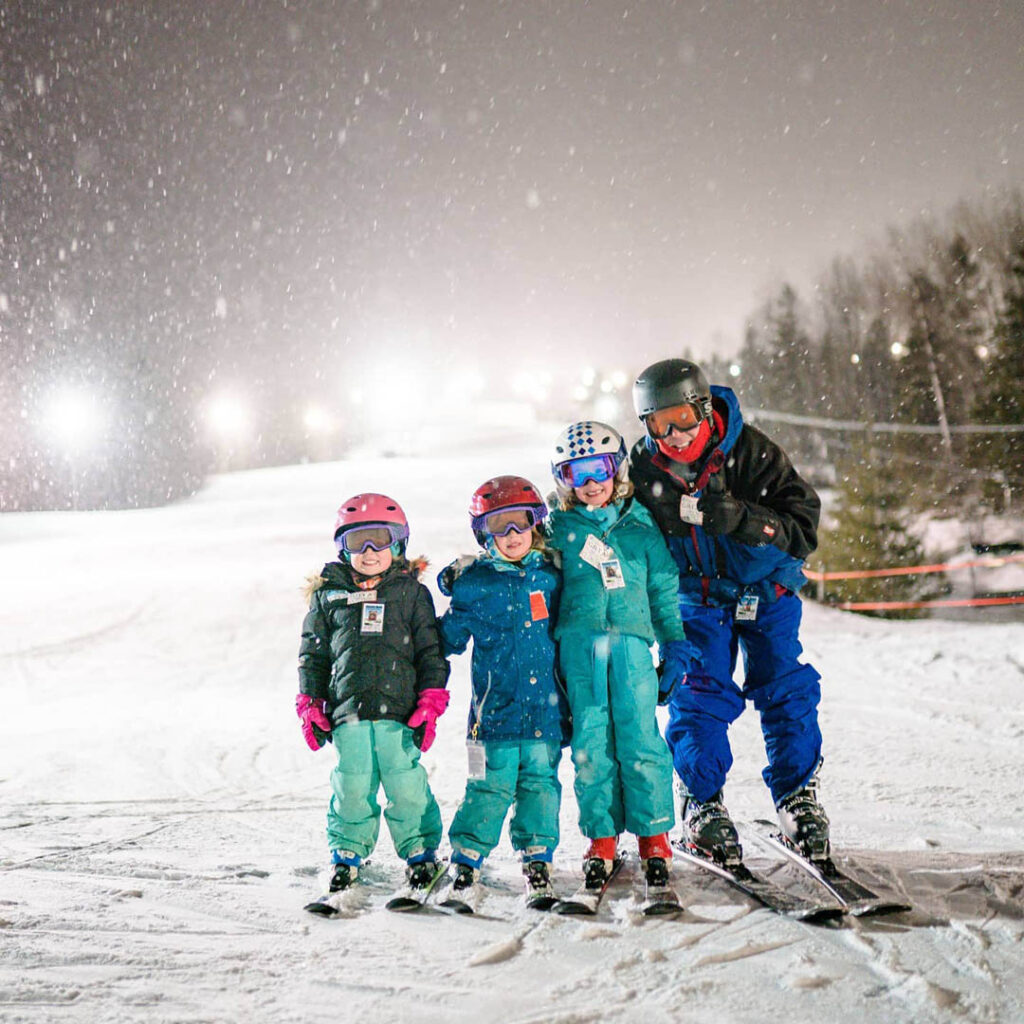 Four kids on skis in colorful jackets, snow pants, helmets, pose for camera. There are lights in the distance, it's snowing.