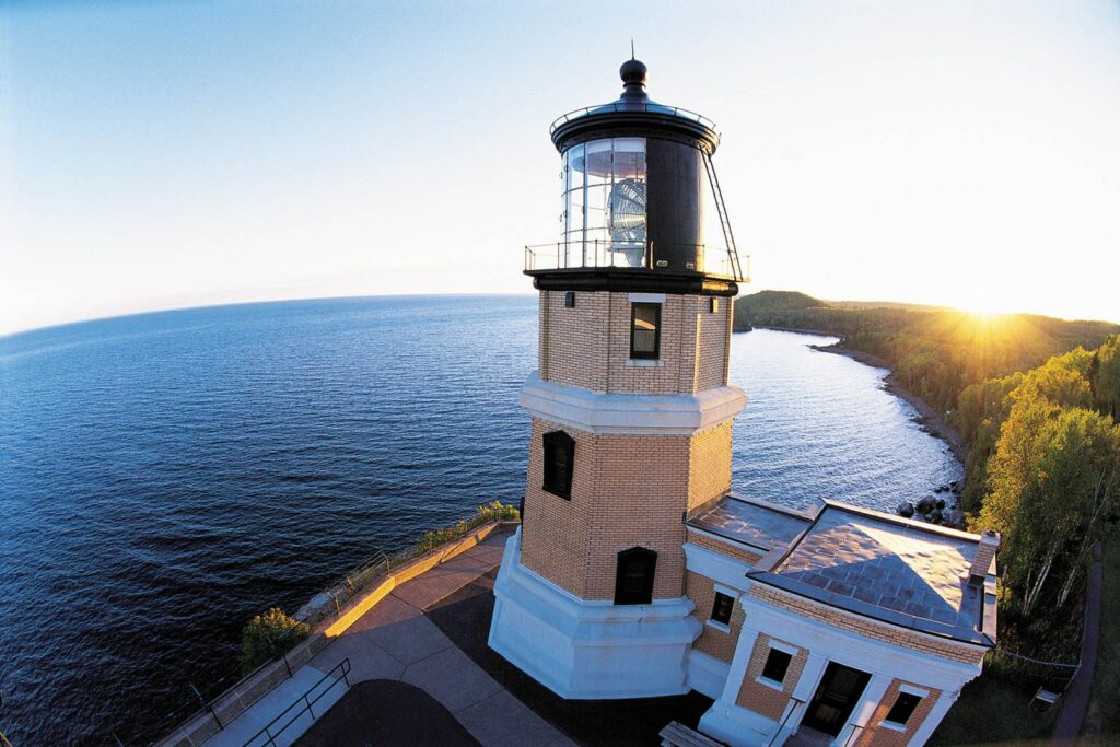 An aerial view of Splitrock Lighthouse with a sunset in the background. The water is very blue.