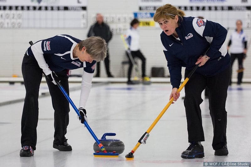 Two women curlers in uniforms sweeping ice. One has a very intense and competitive look on her face.