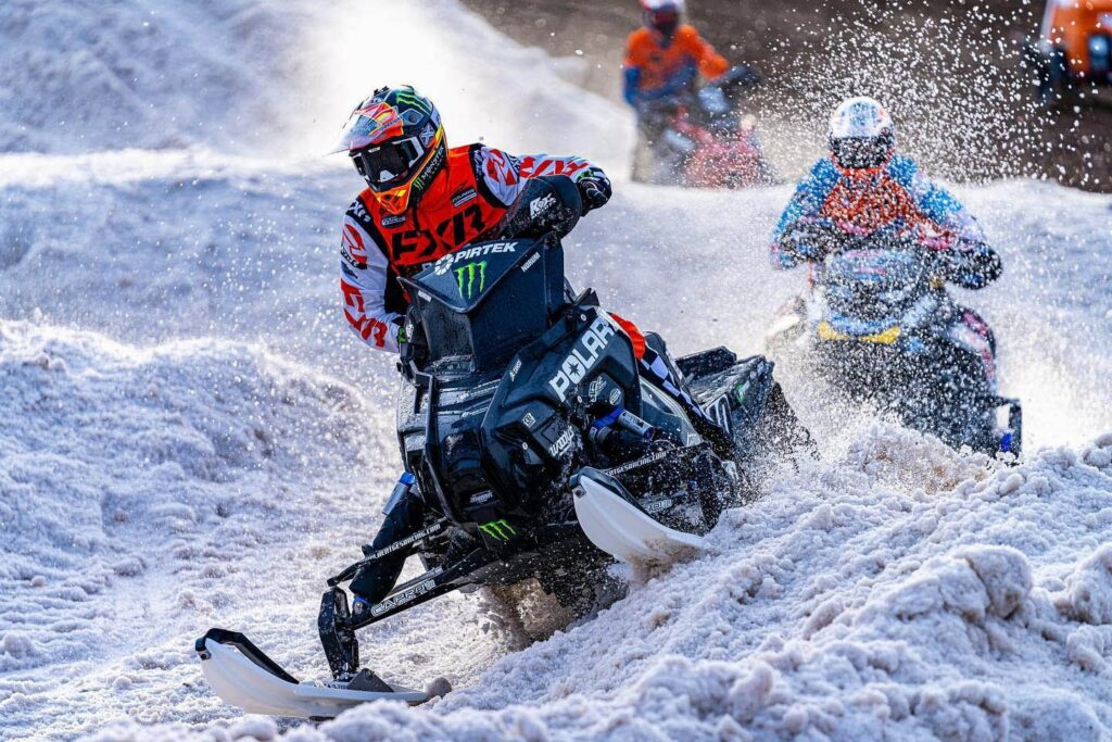 Fast-paced, colorful snowmobilers on machines racing. Snow is spraying everywhere.