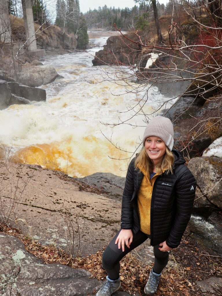 A young woman with blonde hair, wearing a coat and hat is standing next to a very rapid waterfall, smiling at the cabin.