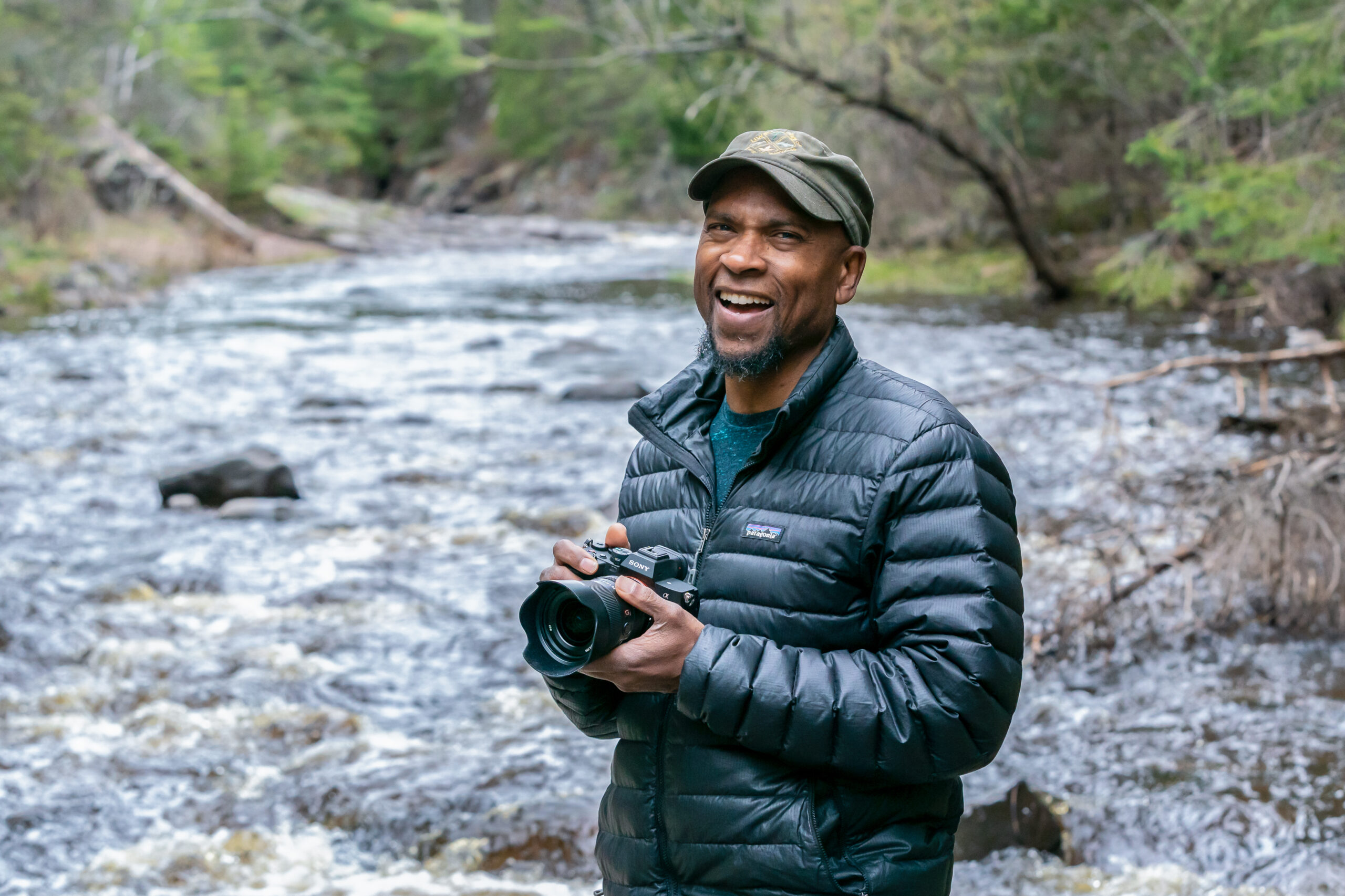 Dudley stands in front of a river smiling with his camera.