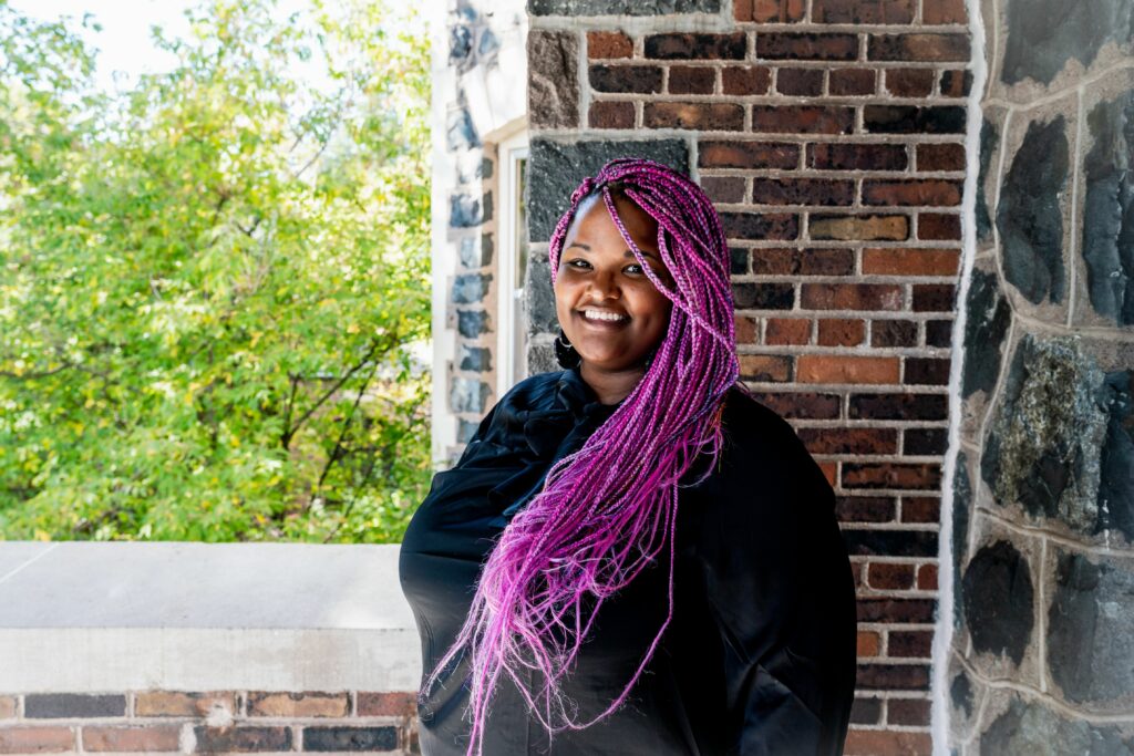 Diona smiles in front of a window. She has purple braids. 