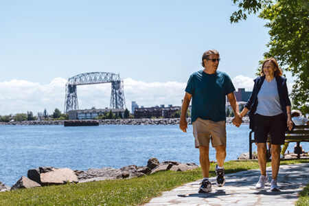 A couple holds hands as they walk on the lakewalk in duluth, mn with the aerial lift bridge and lake superior in the background