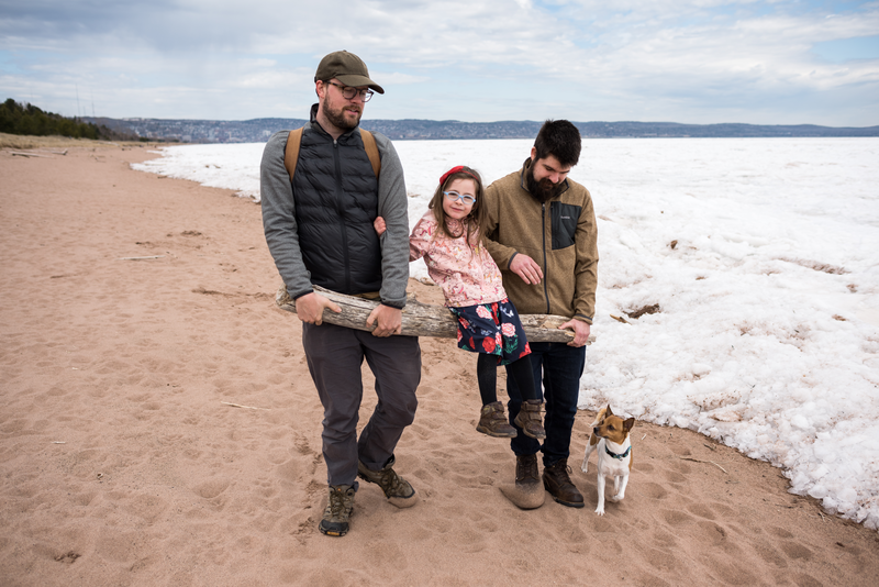A family and their dog walk along the shore of Lake Superior, carrying a child 