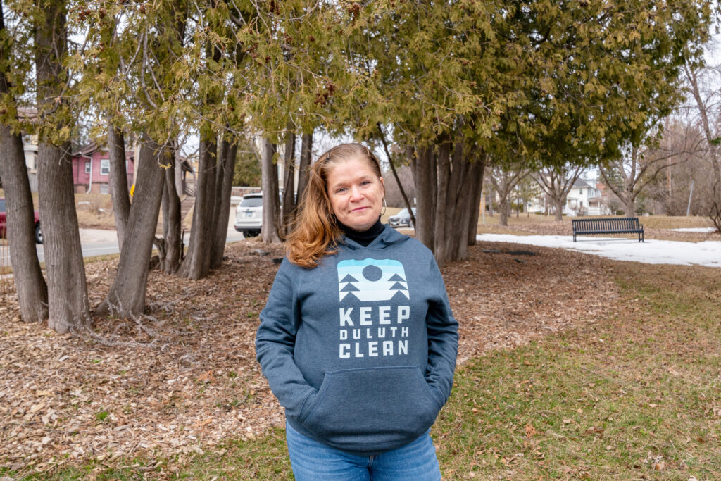 Cheryl Skafte stands outside in Duluth in front of trees, with her hands in the pocket of a Keep Duluth Clean hooded sweatshirt