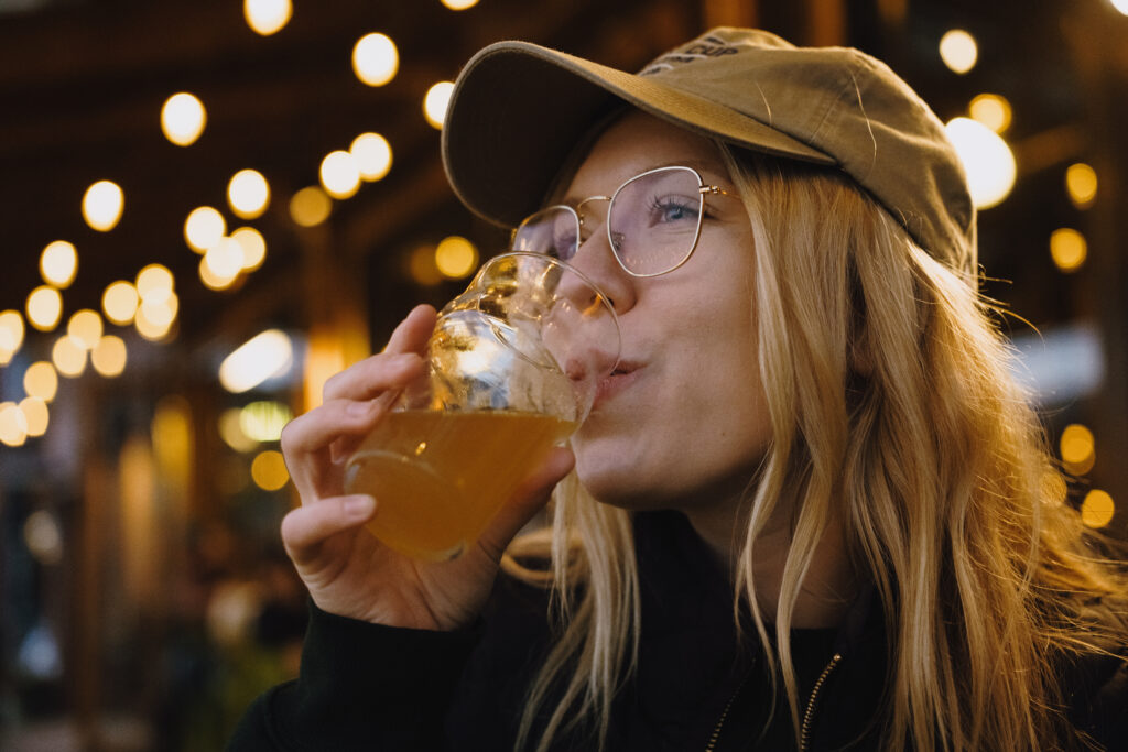 woman wearing a hat drinking a glass of beer with twinkle lights behind her