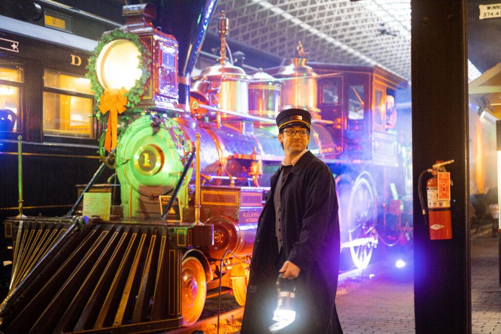 man dressed as a railroad conductor walking in front of a stationary train that is colorfully lit up