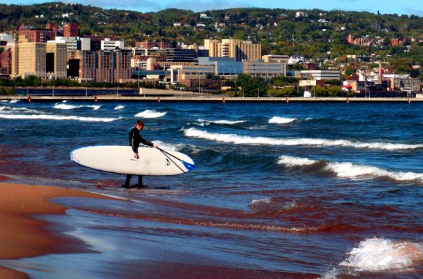 A person wearing a wetsuit carries a surfboard into lake superior in duluth, mn