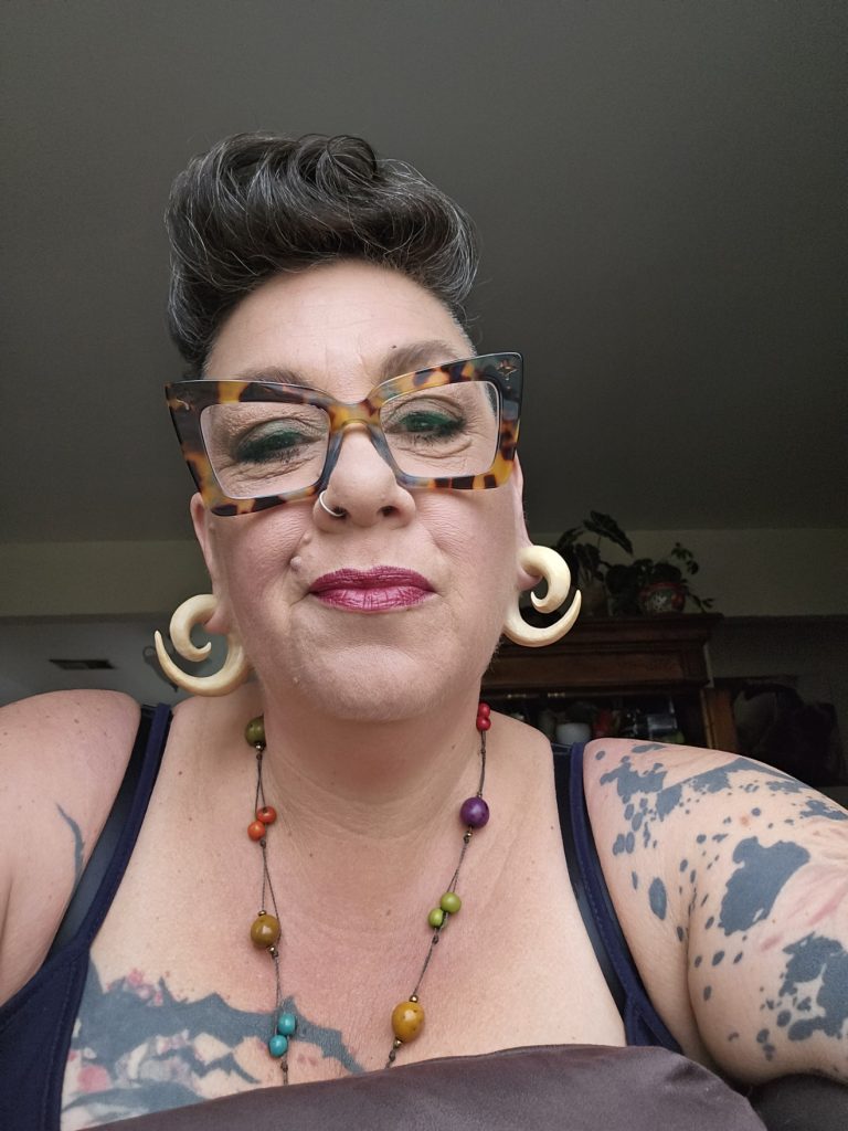 Duluth-based kristy johnson wears a tank top, gold hoops, and a beaded necklace, and large glasses while showcasing her tattoos.