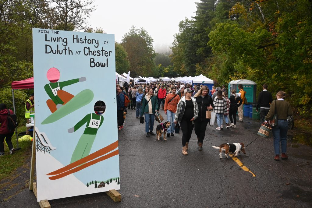 Chester bowl fall fest returns to duluth 2 A ski and snowboard photo cutout board reading, living history of duluth at chester bowl, next to festival attendees walking along vendor-lined streets at fall fest.