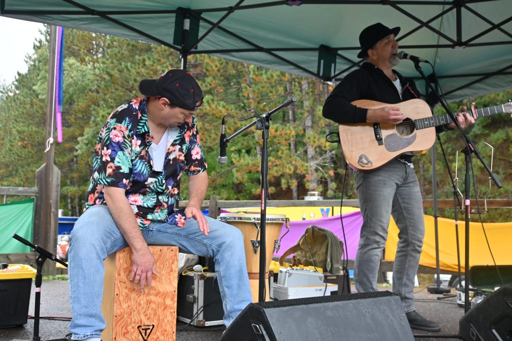 Chester bowl fall fest returns to duluth 3 Two musicians performing live outdoors under a canopy stage at chester bowl fall fest, with one playing acoustic guitar and singing while the other plays a cajón box drum.