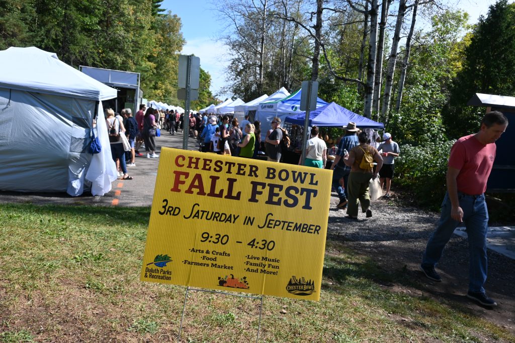 Chester bowl fall fest returns to duluth 1 Chester bowl fall fest yellow event sign on a lawn with crowds of people walking past craft vendor tents in duluth, minnesota.