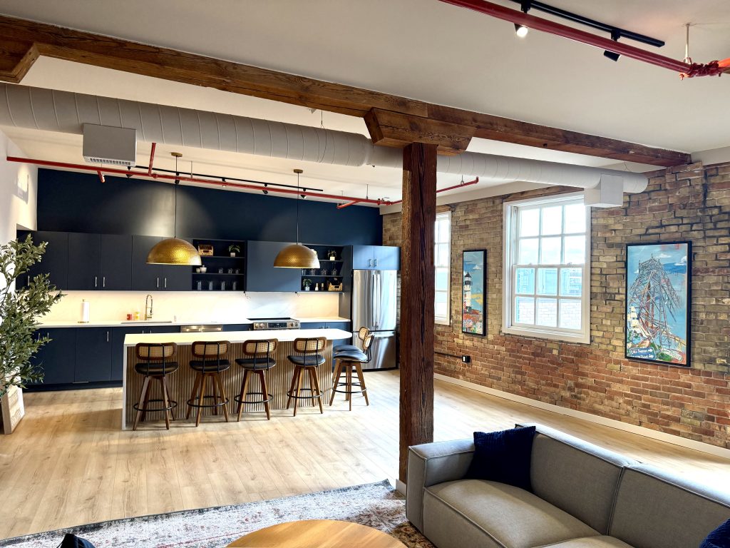 The brix: a different kind of stay in duluth 3 Open-concept loft living space featuring exposed brick walls, timber beams, navy kitchen cabinets, and a fluted island with bar seating.