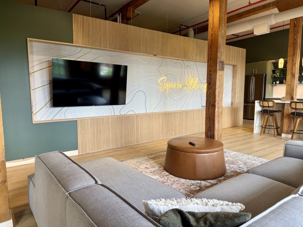 The brix: a different kind of stay in duluth 4 Modern condo living room featuring a mounted tv on a custom line-art feature wall with a glowing superior state of mind neon sign, gray sectional sofa, and leather ottoman.