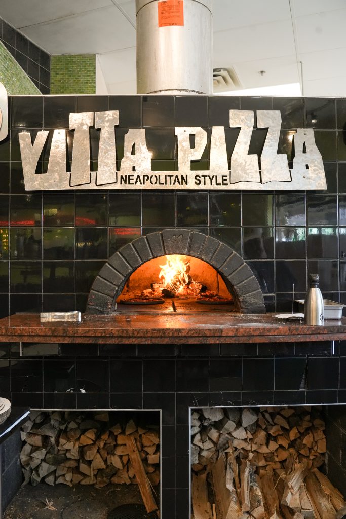 How vitta pizza became a duluth favorite 5 Black tiled wood-fired pizza oven at vitta pizza in duluth with open flames, stacked firewood beneath, and a metallic vitta pizza neapolitan style sign.