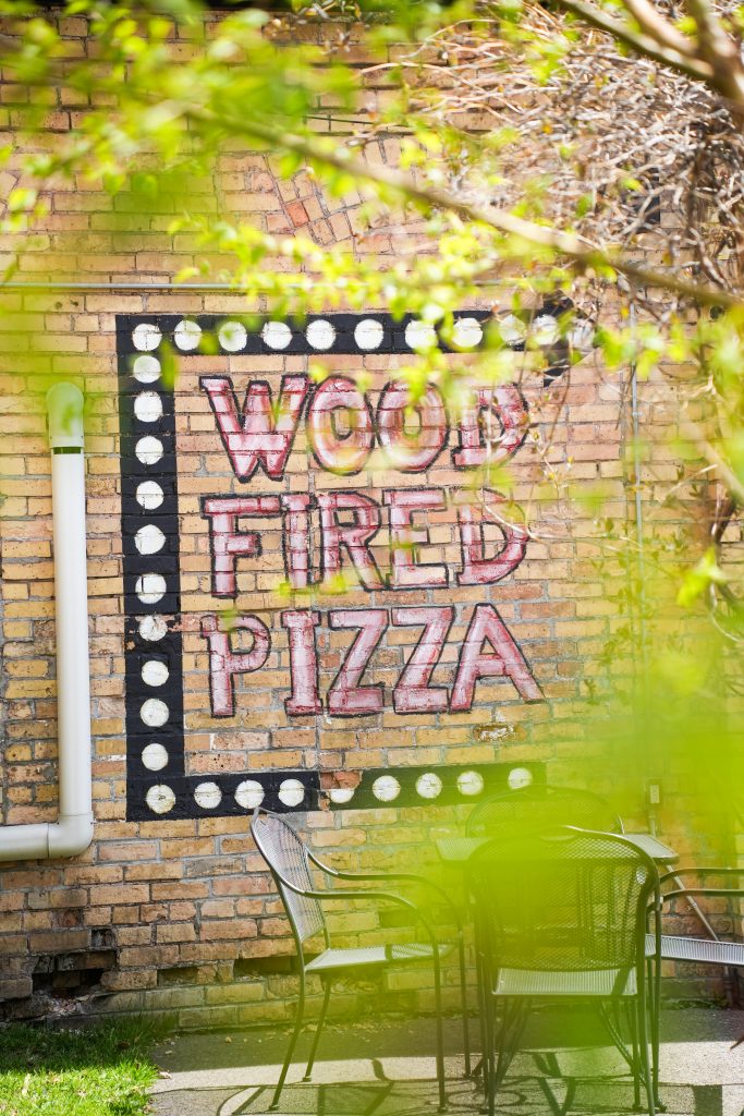 How vitta pizza became a duluth favorite 13 Outdoor patio seating framed by green leaves, with a painted brick wall sign reading wood fired pizza at vitta pizza in duluth.