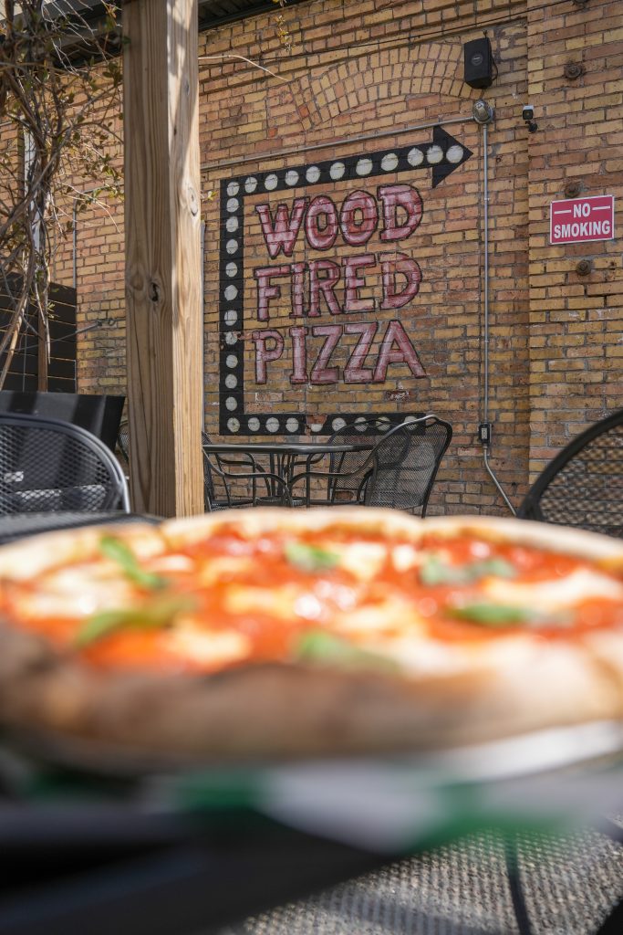 How vitta pizza became a duluth favorite 6 Outdoor patio dining area at vitta pizza in duluth with a painted brick wall sign reading wood fired pizza behind a fresh neapolitan pizza in the foreground.